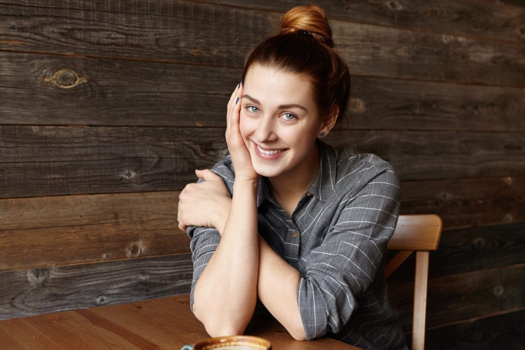 Girl With Bun Smiling While Sitting on Wooden Chair With Elbows on Wooden Table