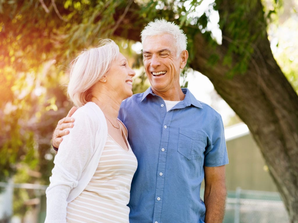 Older Couple Smiling In Front Of Trees With Sun Shining Through